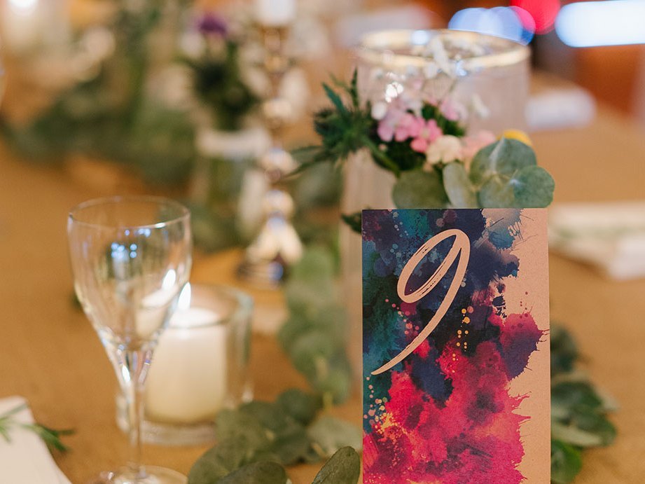 Auf dem Bild ist Deko zu erkennen, die auf einem Tisch steht. Es sind viele Blumen und Kerzen zu sehen. Im Vordergrund steht eine Karte mit bunten Farbklecksen, auf der die Zahl "9" steht.
