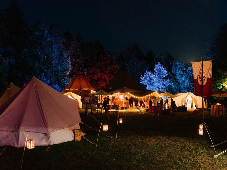 Nachtszenario einer Tipi-Landschaft. Die Bäume im Hintergrund sind bunt beleuchtet, die Zelte strahlen, in warmen Farben.