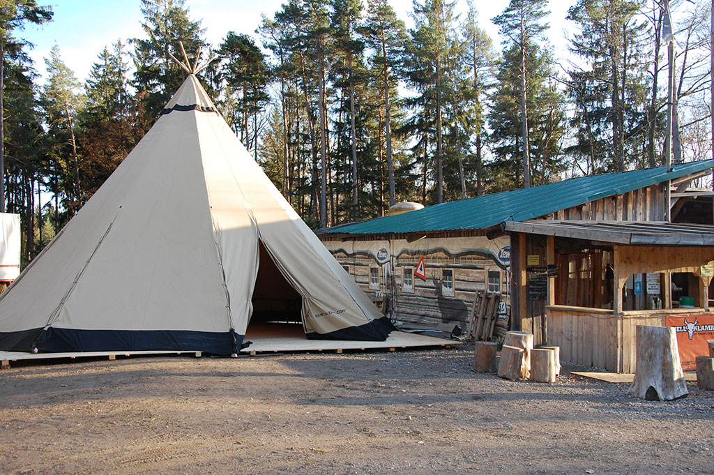 Ein Einzelhut-Tipi steht neben einer Holzhütte. Im Hintergrund ist ein Waldstück zu erkennen.
